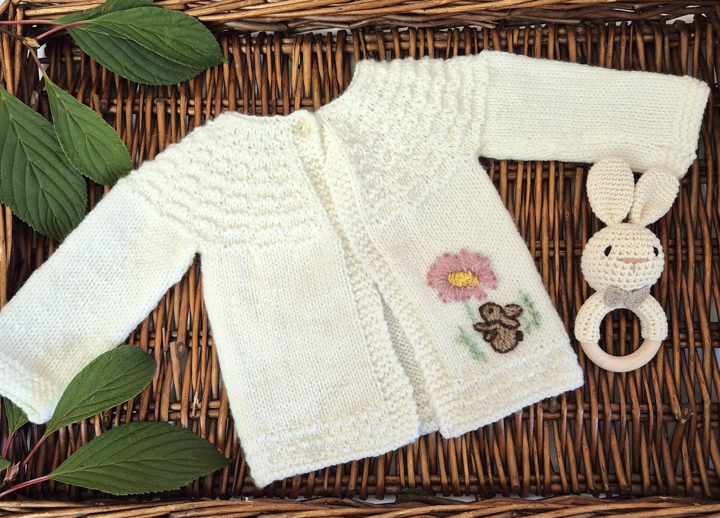 White baby sweater with floral embroidery and a teething ring on a woven surface with leaves.