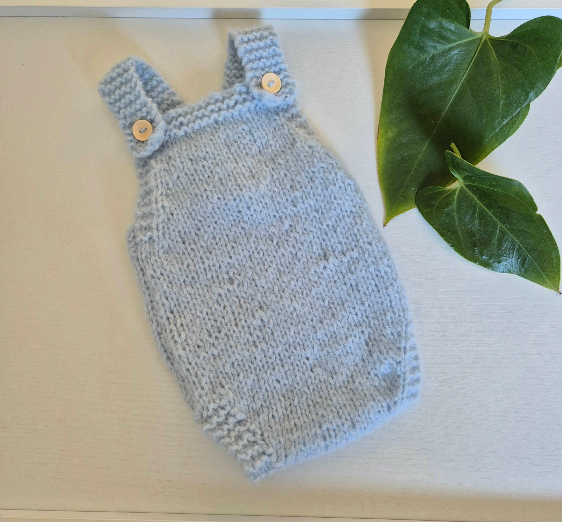 A light blue hand-knit baby romper with buttoned shoulder straps, laid flat on a light cream surface next to green leaves.