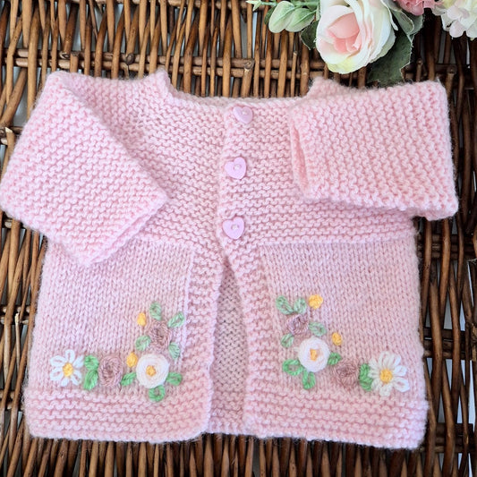 Pink knitted baby cardigan with floral patterns on a wicker surface.