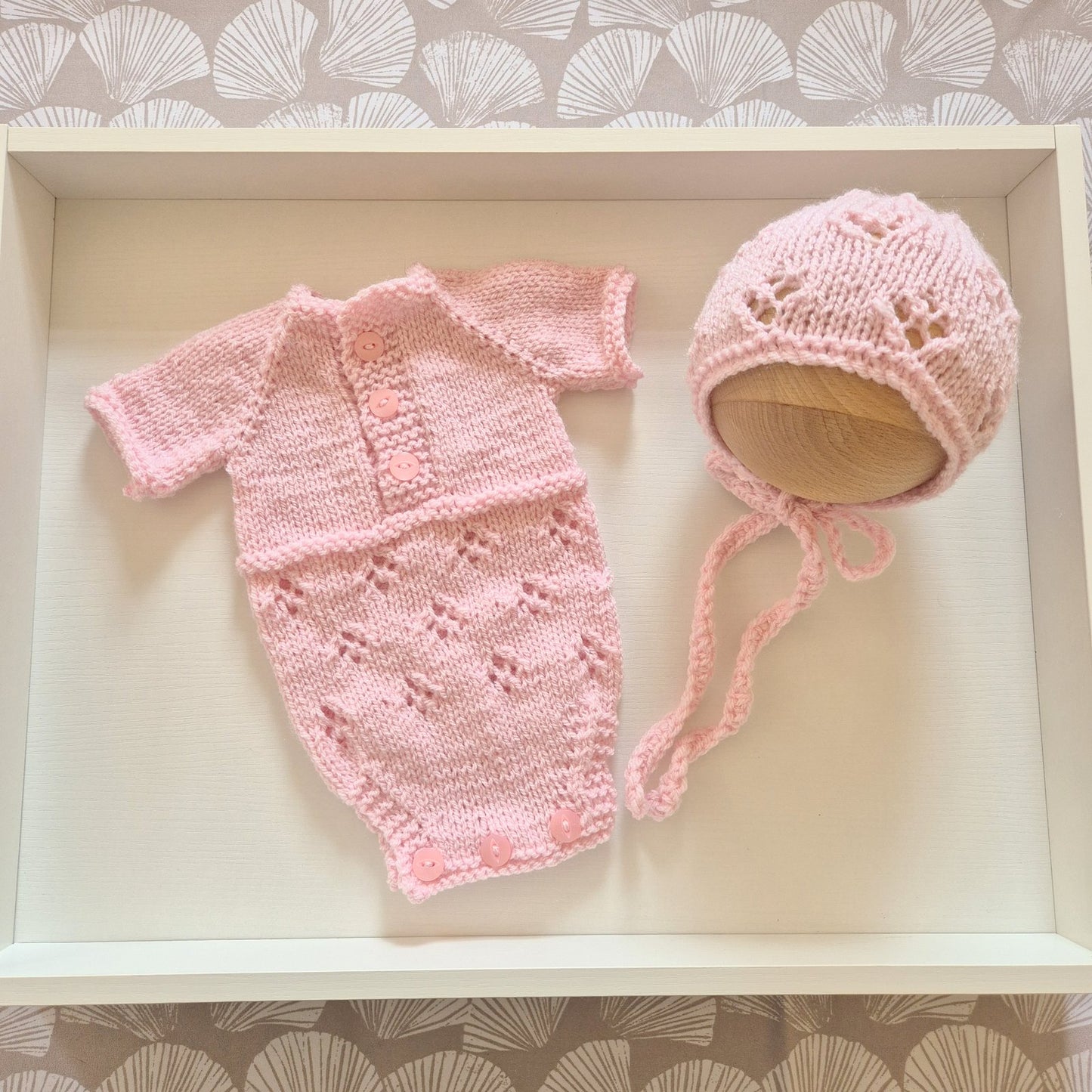 A light pink hand-knit baby set consisting of a short-sleeve romper and a matching bonnet, displayed on a white wooden tray.