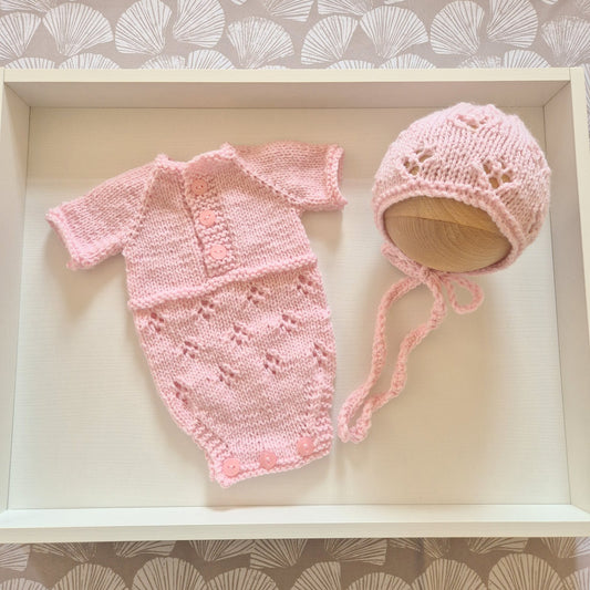 A light pink hand-knit baby set consisting of a short-sleeve romper and a matching bonnet, displayed on a white wooden tray.