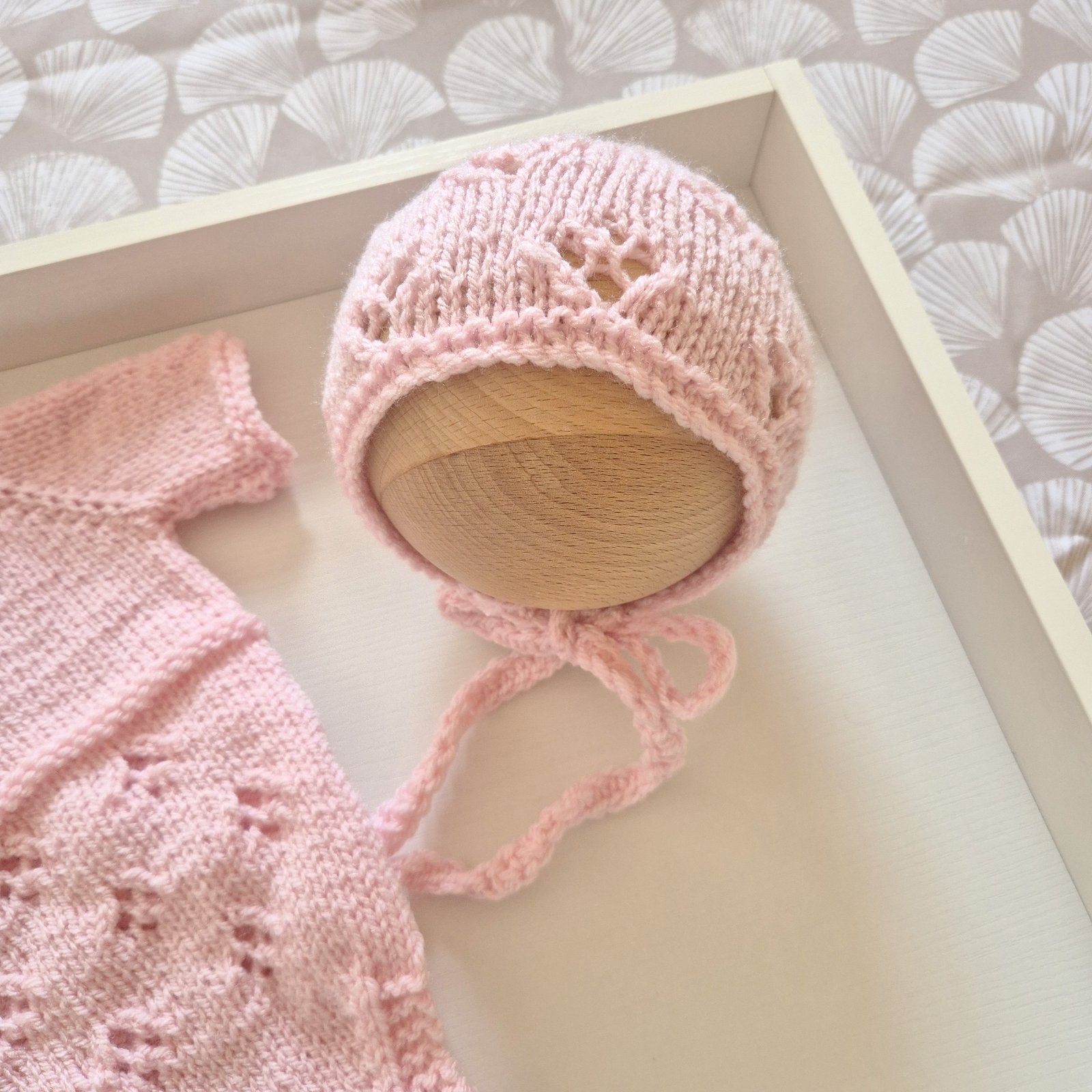 A light pink knitted baby bonnet with lace-like cutout details and tie strings, displayed on a wooden mannequin head inside a white tray. A matching pink knitted garment is partially visible at the left edge.