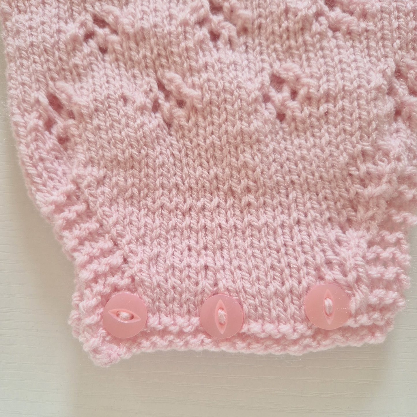 Close-up of a light pink knitted textile with a textured lace-like pattern and three matching pink buttons along the edge, laid on a pale off-white background.
