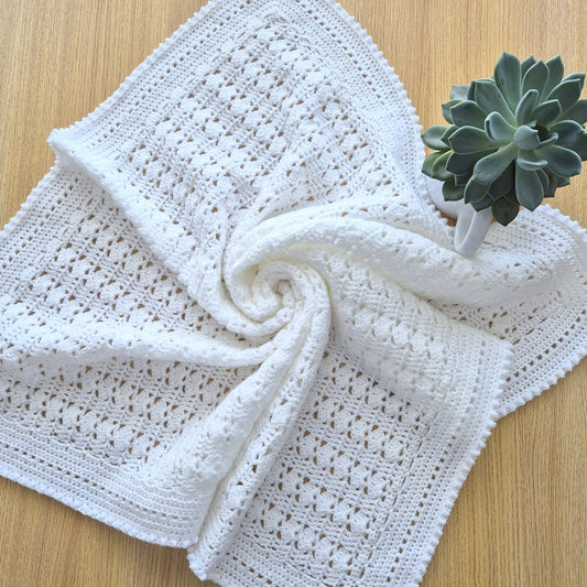 White crocheted blanket on a wooden surface with a small plant.