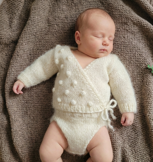 Newborn baby wearing a cream knitted kimono crossover jumper with matching knit pants, on a brown textured blanket