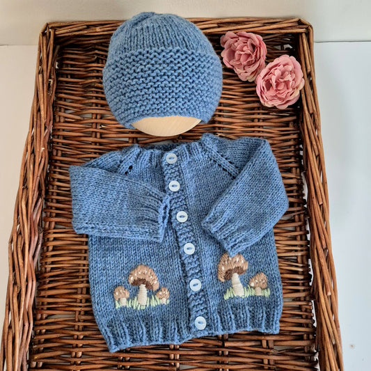 Knitted Dark Blue cardigan and hat set for a baby boy.  The set comprises a gorgeous hand knit baby cardigan with brown embroidered toadstool mushrooms on the front. A matching beanie completes the set. 
Designed to fit a 0-3 month old baby. The cardigan has blue buttons down the front. 