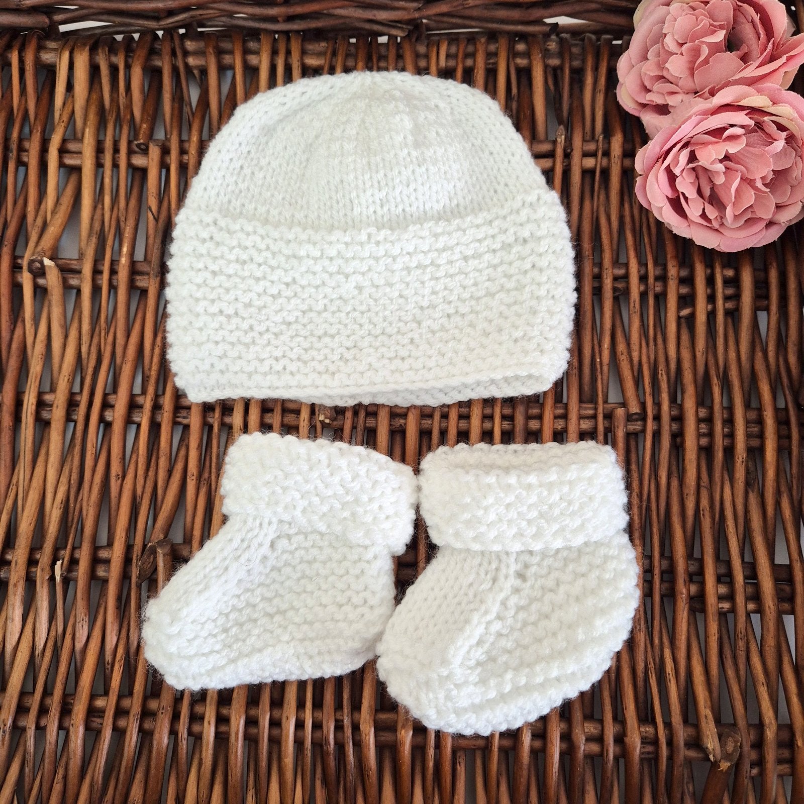 Knitted baby hat and booties on a wicker surface with pink flowers.