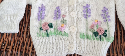 Ready to send handknit white baby cardigan for newborns aged 0–3 months. This charming cardigan features a classic v-neck, front button closure, and delicate embroidered roses, lavender, and flowers across the chest.

Perfect as a baby shower gift or part of a hospital coming home outfit, this one-of-a-kind piece arrives beautifully presented in an organza gift bag.