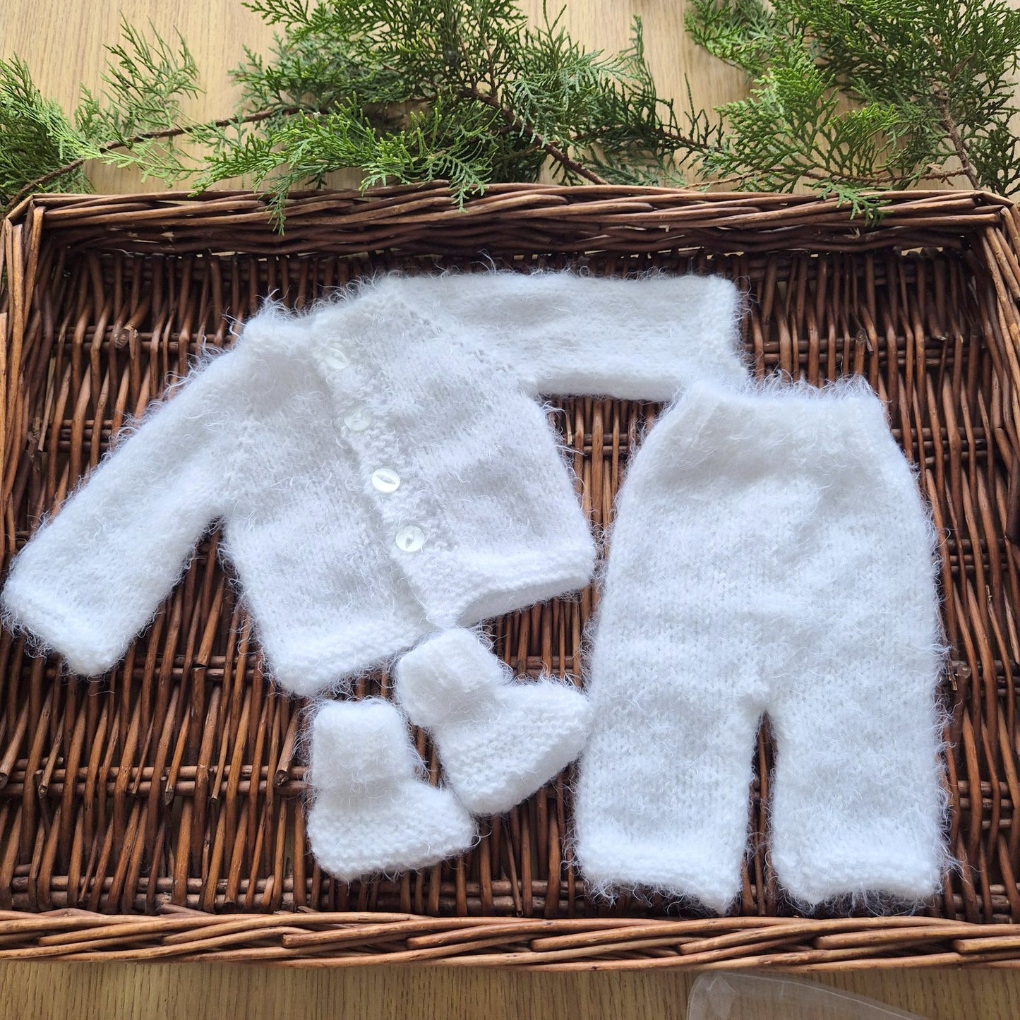 White baby outfit with jacket, pants, and booties on a woven basket with greenery.
