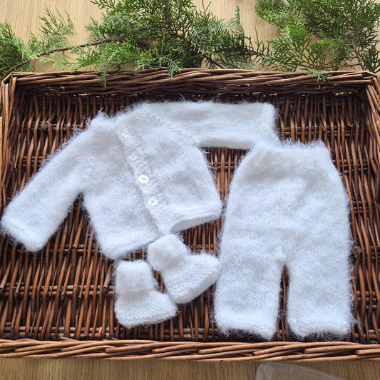 White baby outfit with jacket, pants, and booties on a woven basket with greenery.
