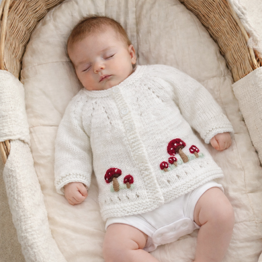 Baby wearing a white knitted cardigan with mushroom patterns, lying in a basket.