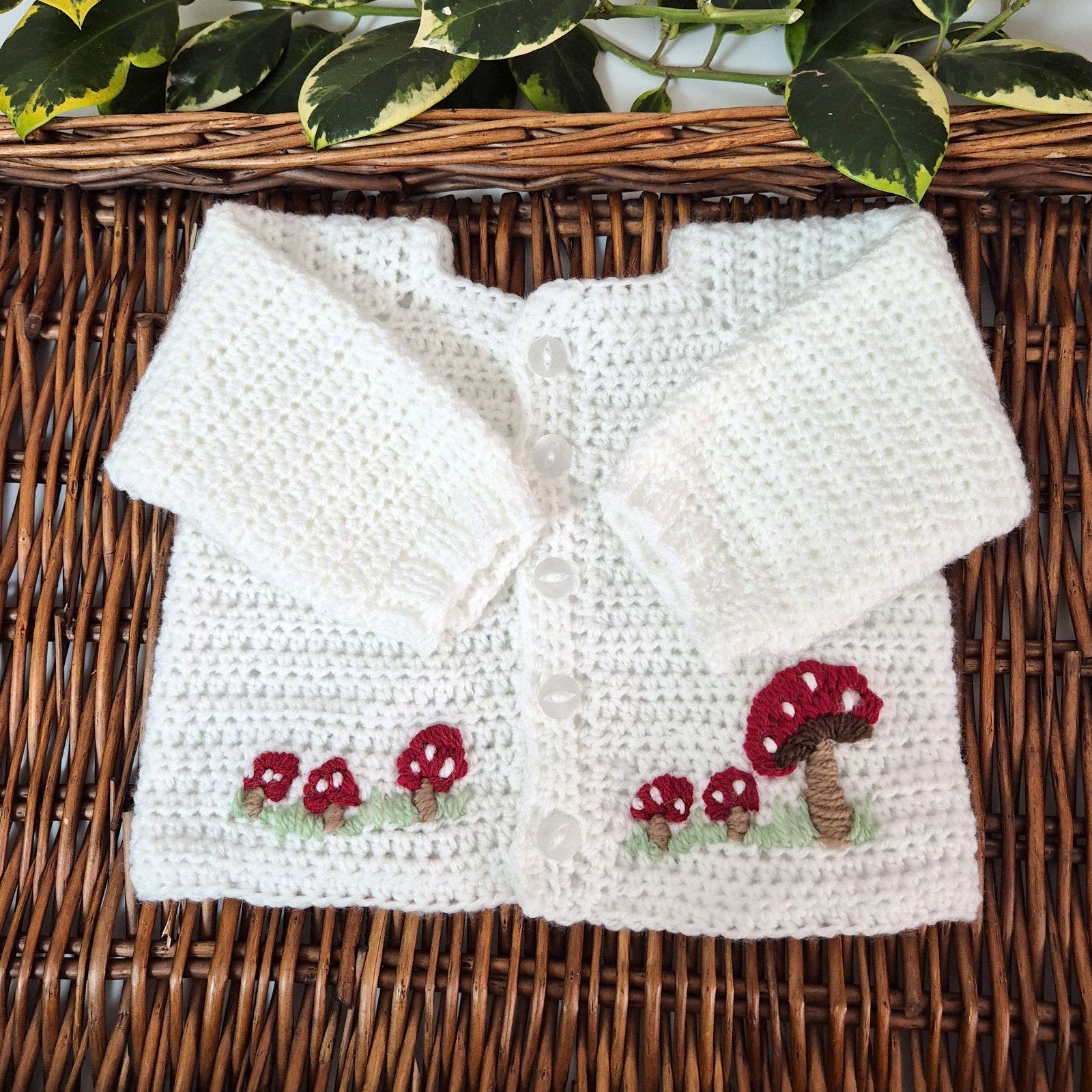White crocheted baby cardigan with mushroom designs on a wicker surface.