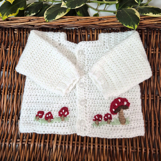 White crocheted baby cardigan with mushroom designs on a wicker surface.