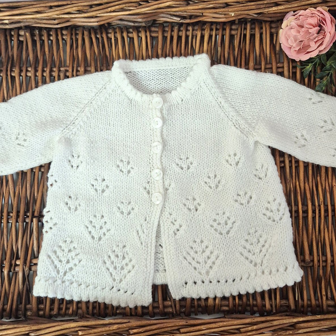 White knitted baby cardigan on a woven surface with pink flowers