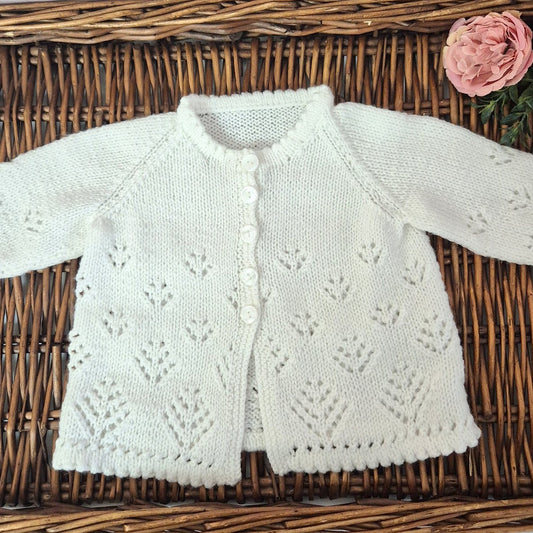 White knitted baby cardigan on a woven surface with pink flowers