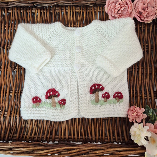 White knitted cardigan with mushroom design on a wicker surface with pink flowers.
